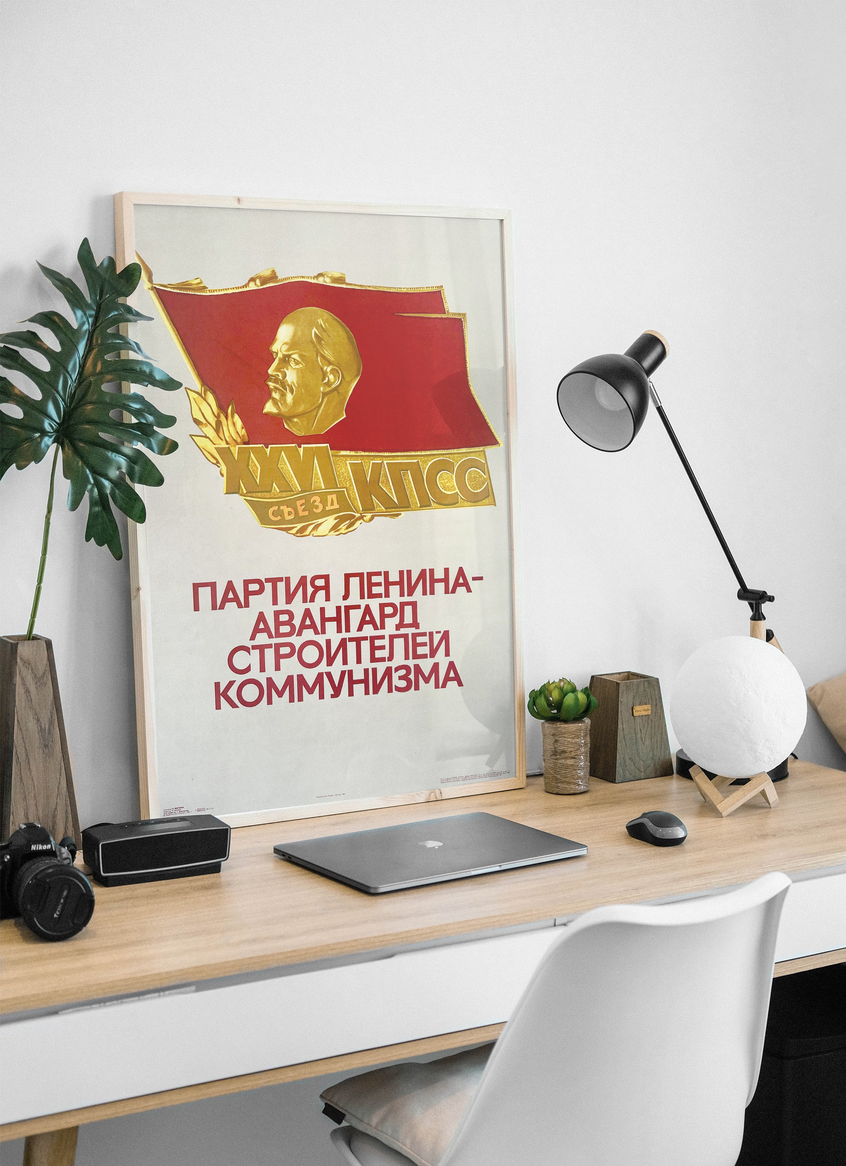 A framed Soviet-style poster featuring a red flag with a gold portrait and Cyrillic text, displayed on a light wood desk in a modern home office setup with a laptop, lamp, and plant.