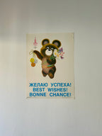 Poster with Moscow Olympic bear holding flowers, text in Russian, English, and French on a white wall.