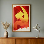 Framed poster with a yellow hammer and sickle symbol on a red background, placed on a wooden shelf against a gray wall.