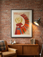 Framed vintage poster on a brick wall with a wooden cabinet and chair.