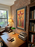 Home office with a desk, lamp, books, and a framed poster on a brick wall.