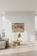 A modern living room with a light beige sofa, a round wooden coffee table, and a vintage-style Russian poster hanging on a white wall. The room features large floor-to-ceiling windows with a view of a city skyline, bright natural light, and light wood flooring.
