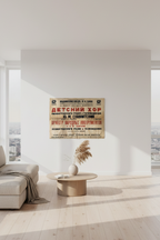 A modern living room with a light beige sofa, a round wooden coffee table, and a vintage-style Russian poster hanging on a white wall. The room features large floor-to-ceiling windows with a view of a city skyline, bright natural light, and light wood flooring.