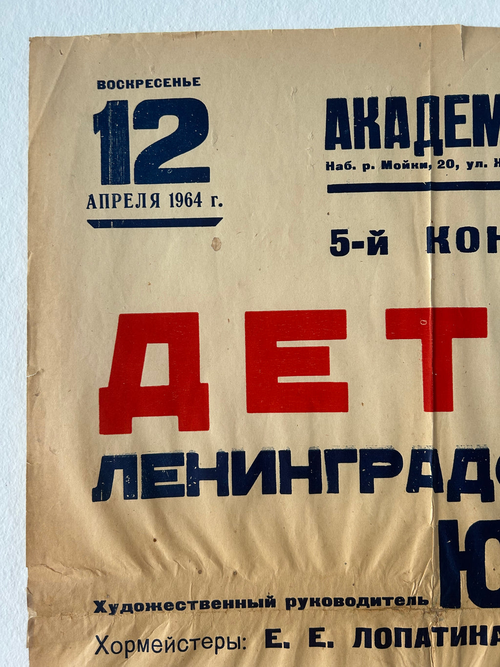 A vintage paper poster or sign with Cyrillic text, featuring large red and dark blue lettering on a beige background, photographed against a white wall.