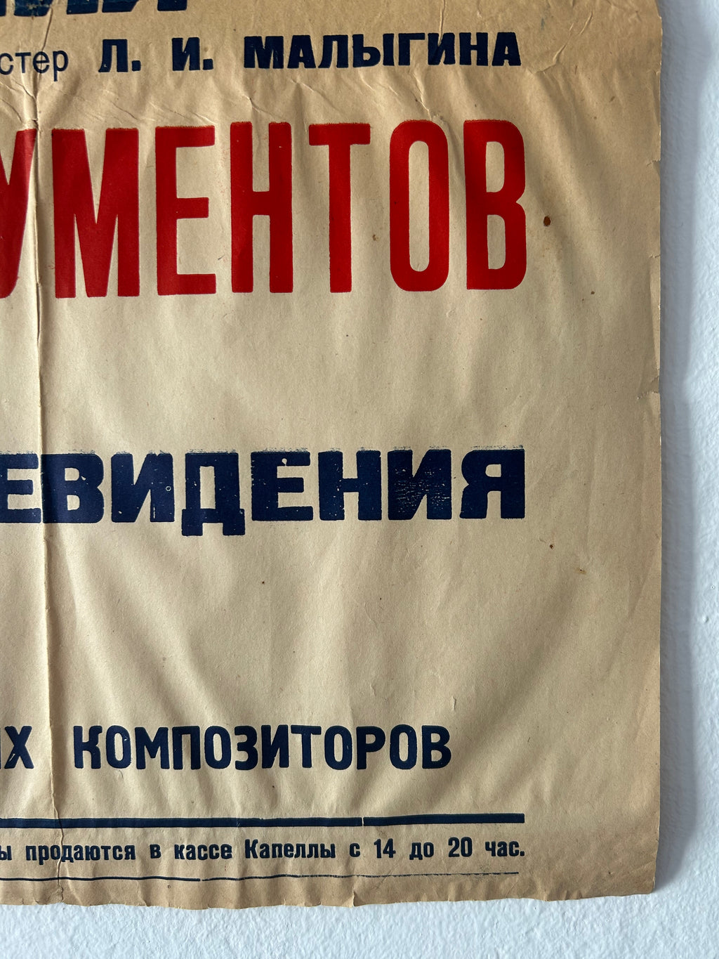 A vintage paper poster in Russian, printed on aged beige paper with bold red and dark blue text, photographed against a white wall.