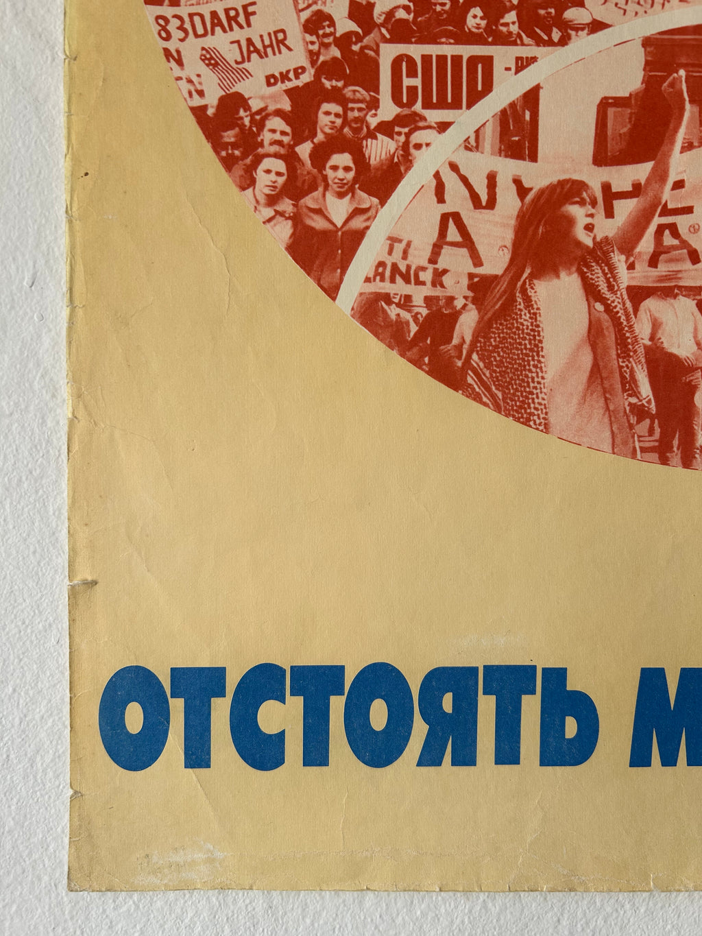 A close-up of a vintage-style poster or printed panel with a beige background, blue vertical Cyrillic text on the right, and a red circular collage of a crowd scene on the left.