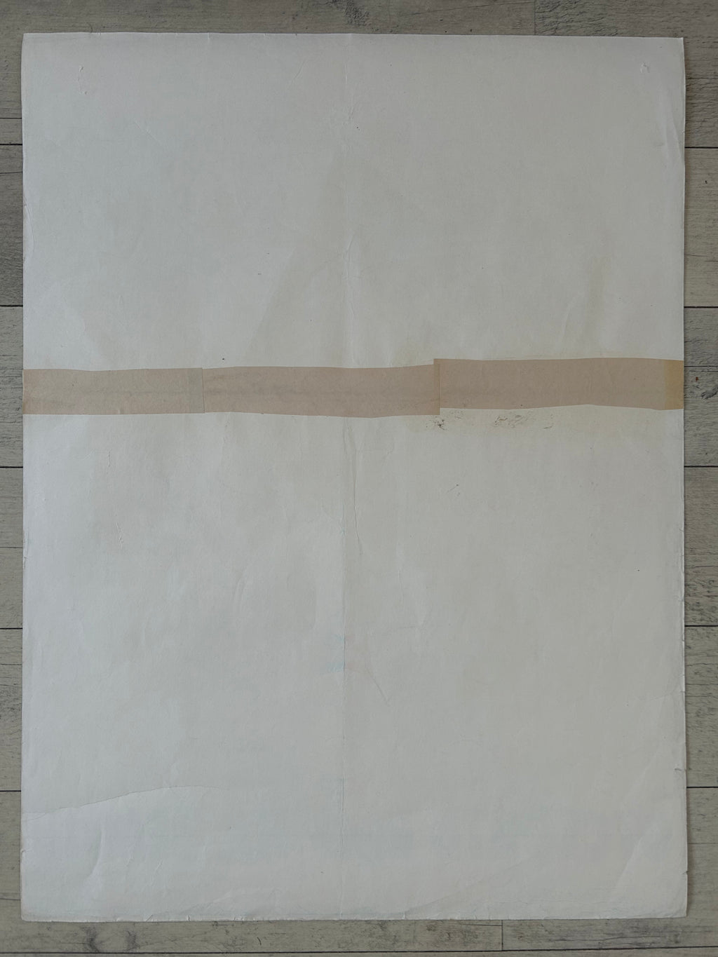A large off-white sheet of paper or fabric laid flat on a gray wood floor, with a narrow beige tape strip crossing horizontally across the middle.