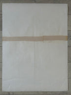 A large off-white sheet of paper or fabric laid flat on a gray wood floor, with a narrow beige tape strip crossing horizontally across the middle.