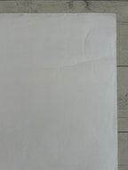 A close-up of a plain white textured surface, likely a sheet of paper, cardstock, or craft foam, laid flat on a light gray wood-grain surface.