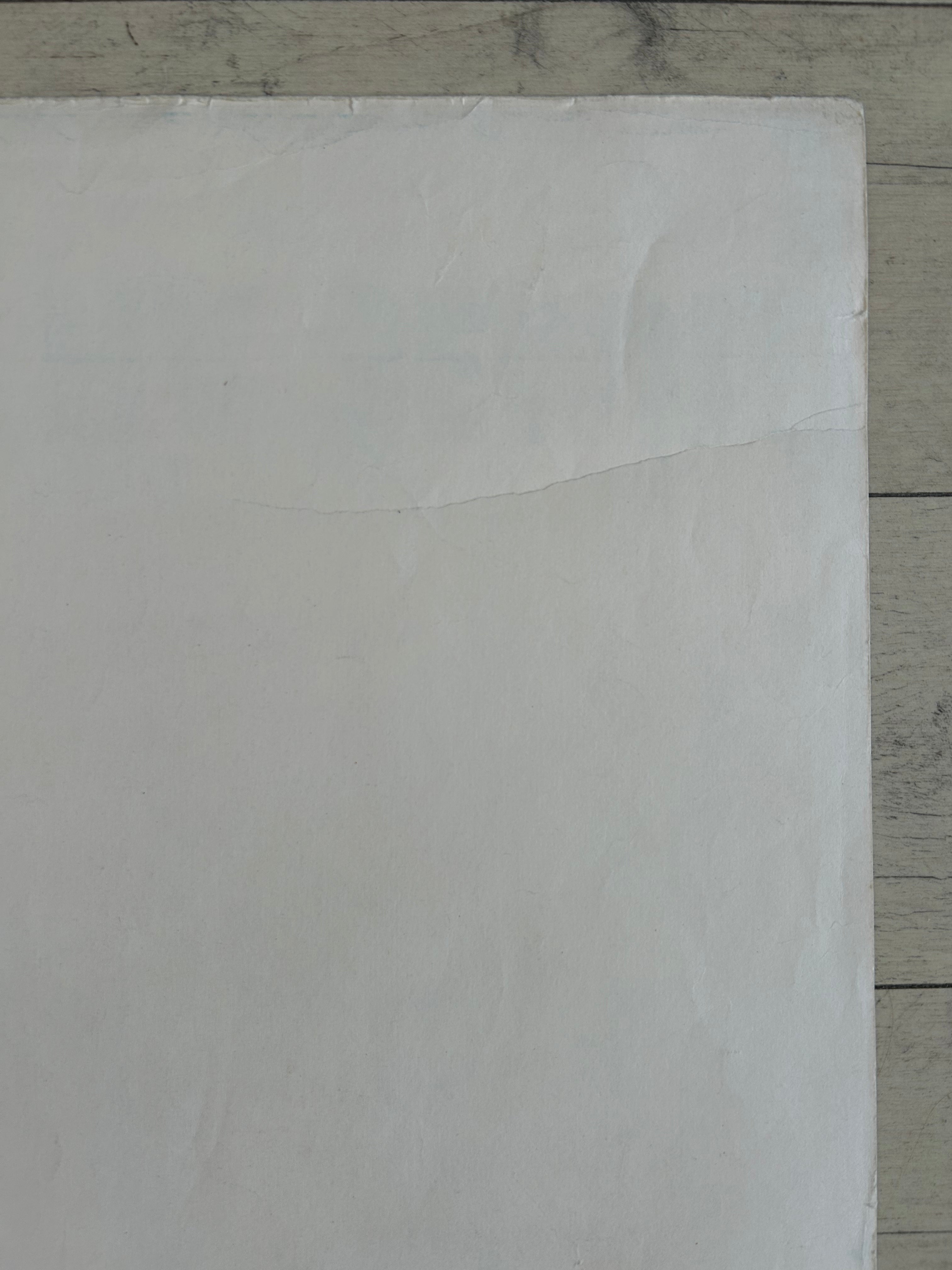 A close-up of a plain white textured surface, likely a sheet of paper, cardstock, or craft foam, laid flat on a light gray wood-grain surface.