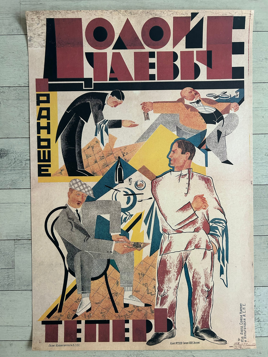 A vintage Soviet propaganda poster print featuring stylized figures in suits and abstract geometric shapes, with large Cyrillic text at the top and bottom, photographed against a light wood background.