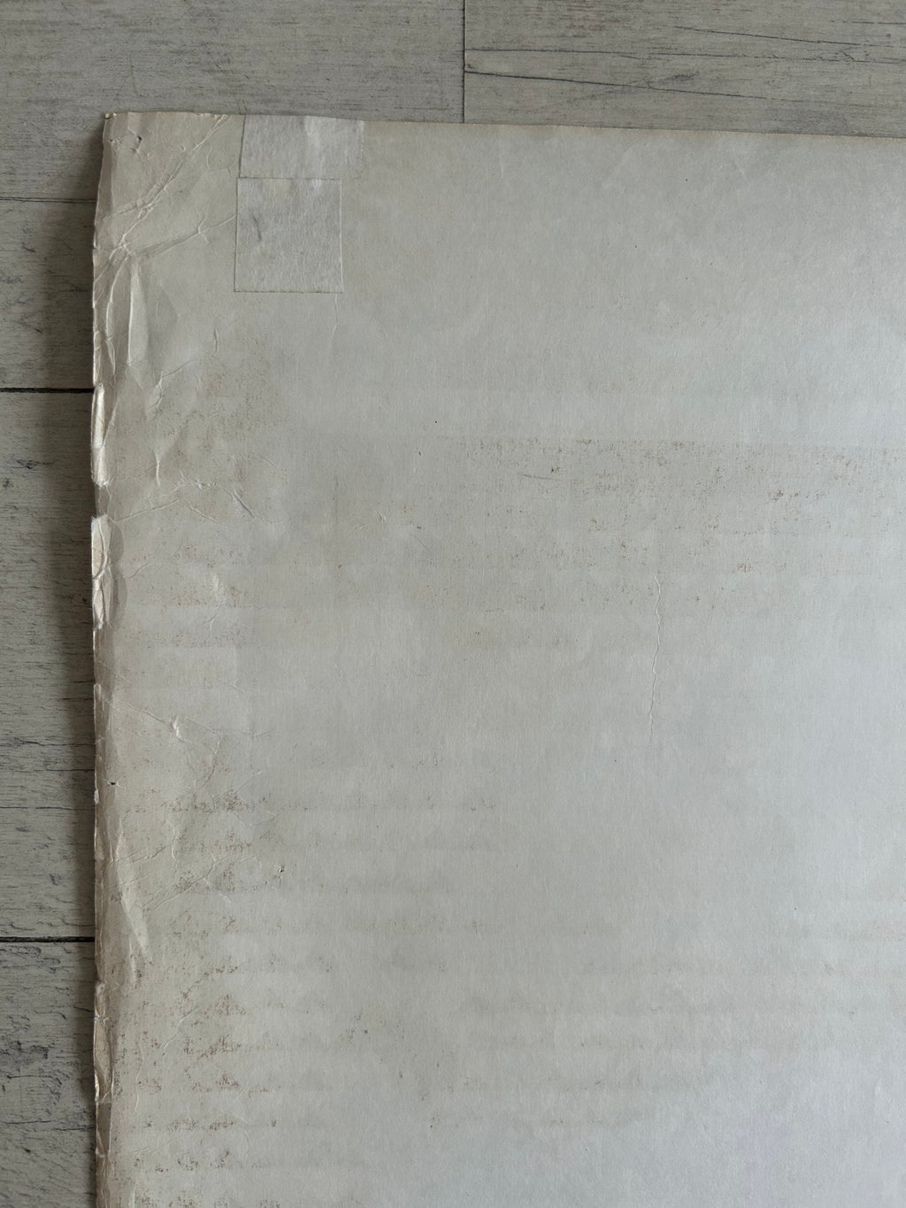A large sheet of off-white textured paper or canvas laid flat on a light gray wood floor, with the top and right edges slightly curled up.