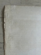 A large sheet of off-white textured paper or canvas laid flat on a light gray wood floor, with the top and right edges slightly curled up.