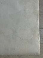 A close-up of a textured, off-white fabric or paper material with a subtle vertical ribbed pattern and faint gray markings, set against a weathered gray wood background.