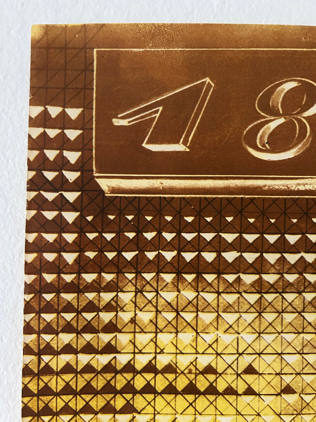 Close-up of a gold-toned patterned surface with geometric triangles and a raised rectangular panel featuring stylized numbers, photographed against a white background.