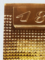 Close-up of a gold-toned patterned surface with geometric triangles and a raised rectangular panel featuring stylized numbers, photographed against a white background.