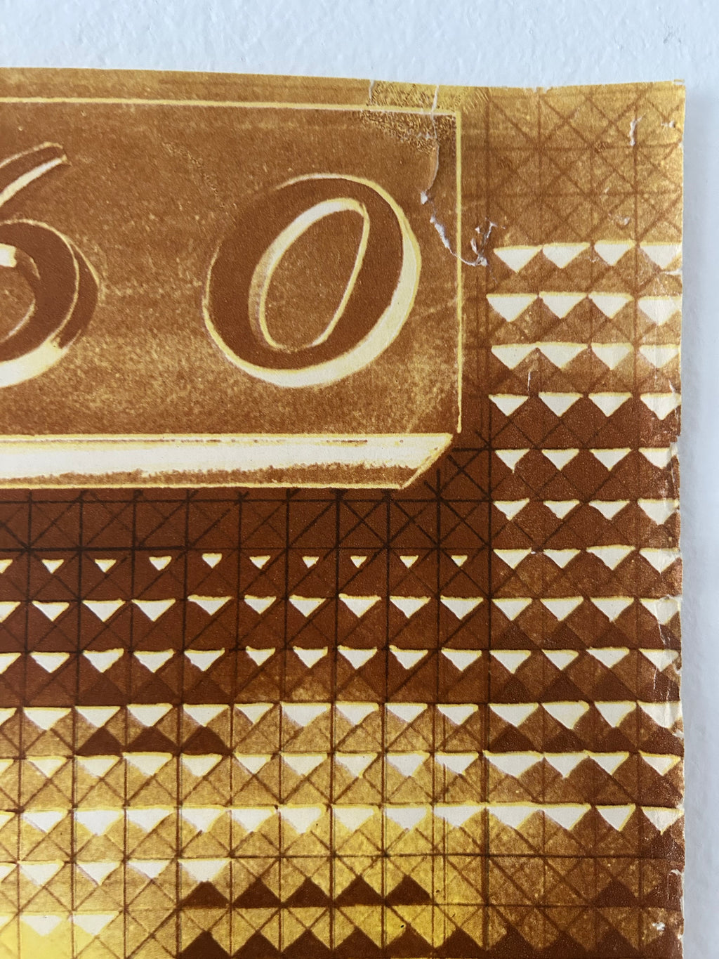 A close-up of a gold-toned patterned surface with repeating geometric triangles and a large circular motif, photographed against a light neutral background.