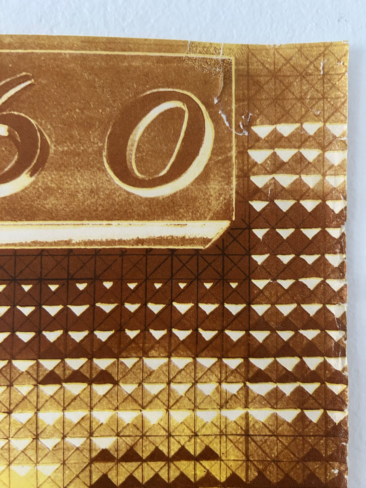 A close-up of a gold-toned patterned surface with repeating geometric triangles and a large circular motif, photographed against a light neutral background.