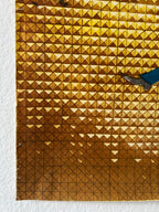 A close-up of a gold-toned geometric textured surface with a triangular pattern, partially bordered by a white textured background and showing a small blue and gray object at the top edge.