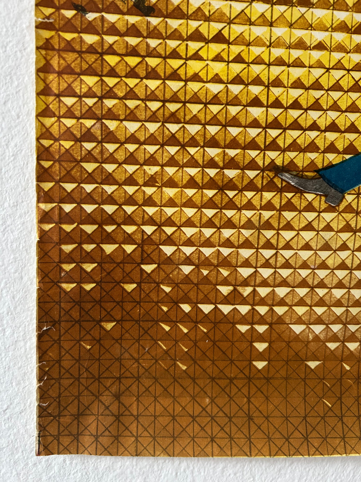 A close-up of a gold-toned geometric textured surface with a triangular pattern, partially bordered by a white textured background and showing a small blue and gray object at the top edge.