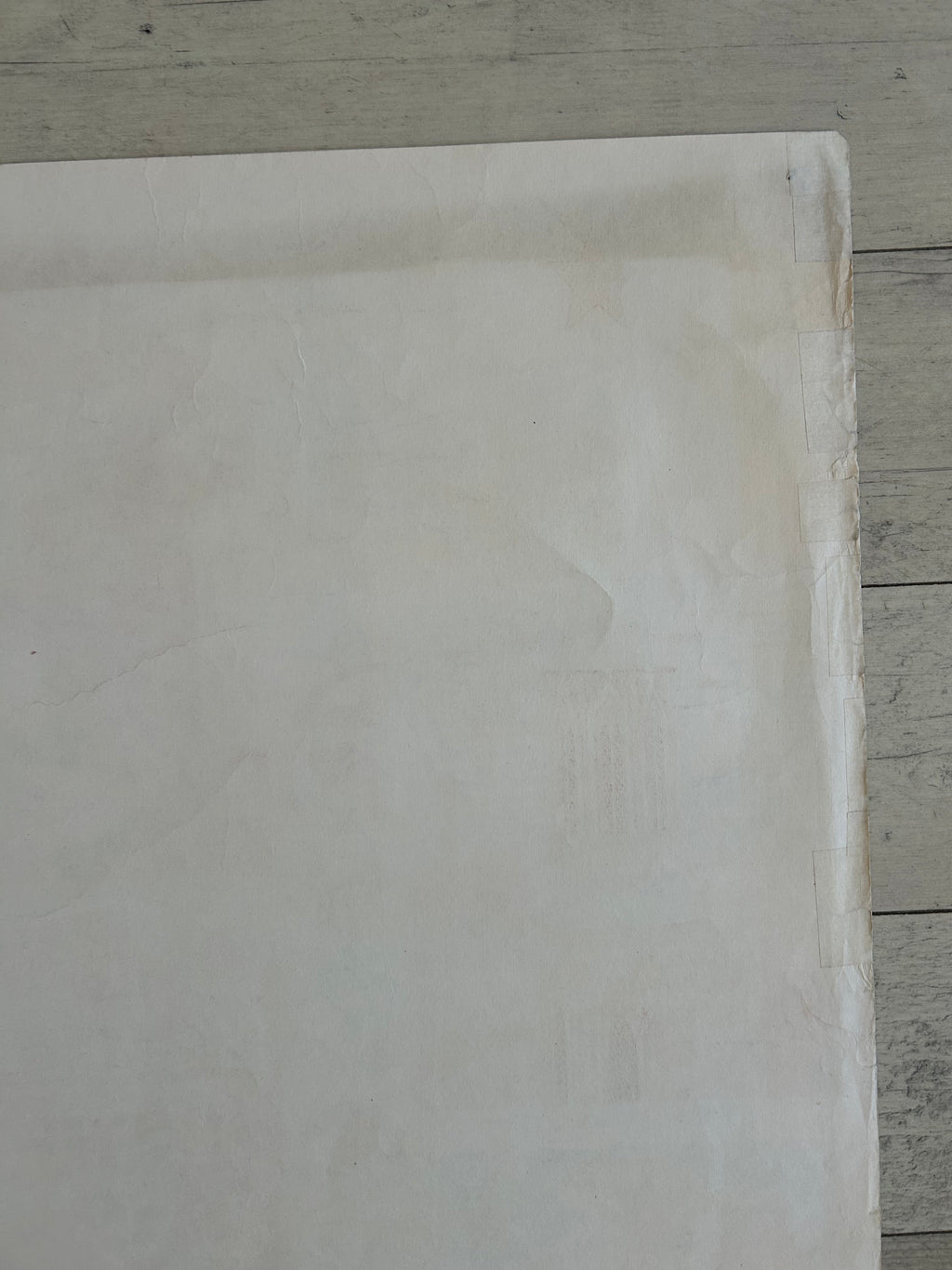 A large, blank off-white square sheet or panel with a slightly textured surface, positioned against a light gray wooden plank background.