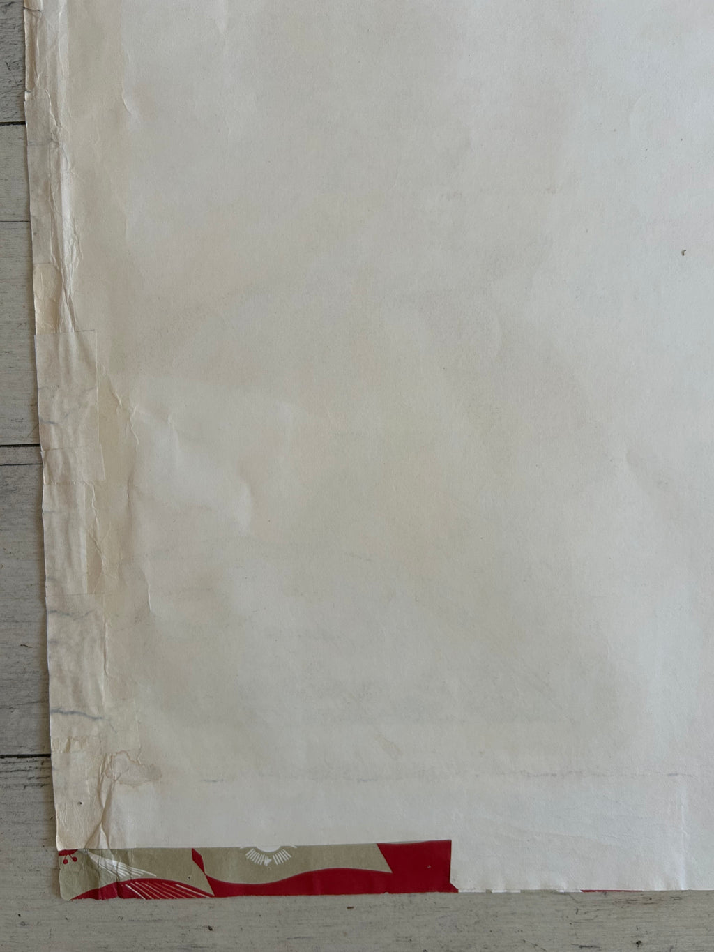 A close-up of a large off-white sheet or panel with visible wrinkles, covering most of the frame, with a small red and beige patterned section visible along the right edge.