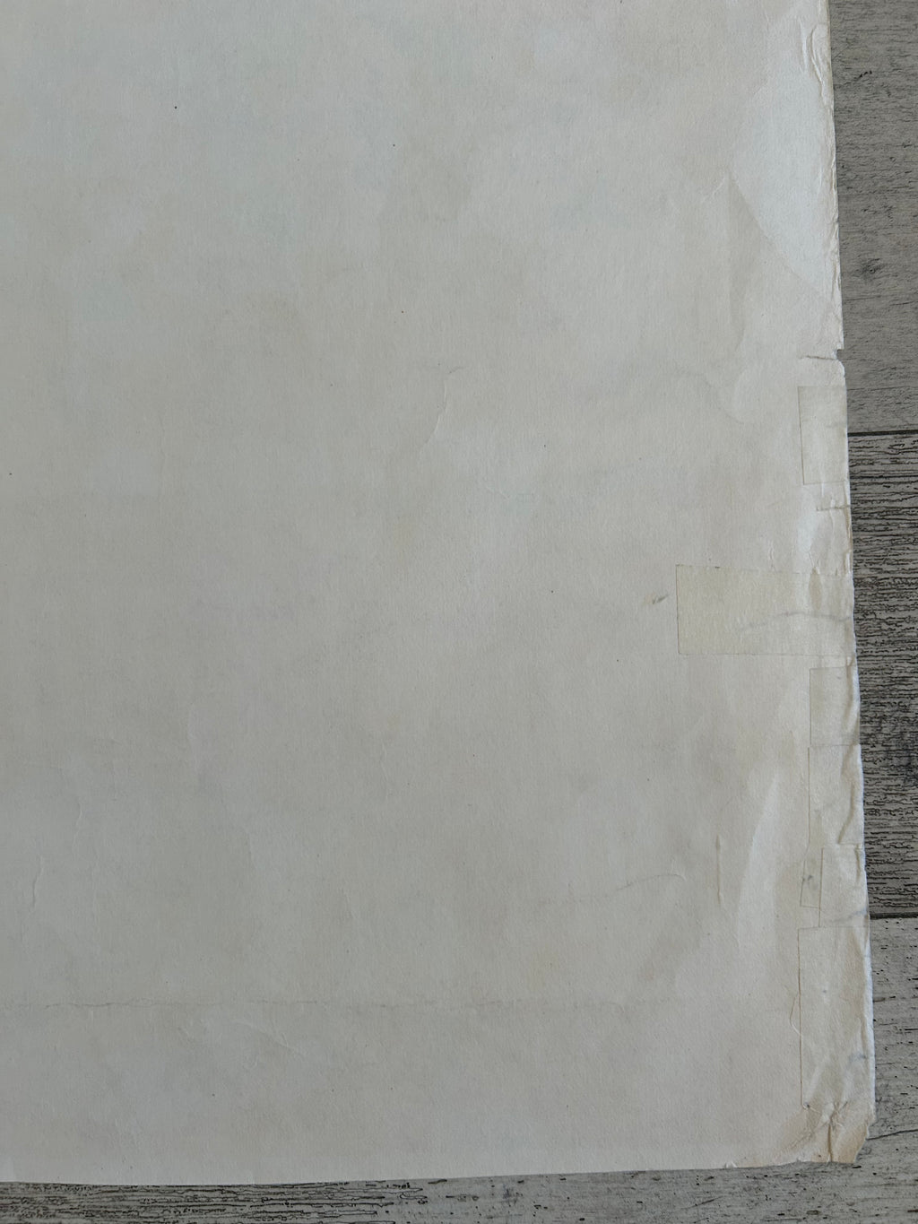A close-up of a large off-white sheet of paper or fabric with a slightly textured surface, taped along one edge, resting on a gray wood surface.