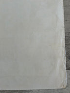 A close-up of a large off-white sheet of paper or fabric with a slightly textured surface, taped along one edge, resting on a gray wood surface.