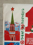 A close-up of a vintage Soviet-style poster or print laid on a light wood floor. The artwork features bold red blocks, green leafy vines, a green spire, blue buildings, and white Cyrillic text.