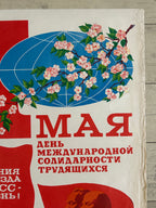 A vintage-style Soviet propaganda poster print featuring a blue globe with pink blossoms, large red Cyrillic text, and a red abstract shape on an off-white background, photographed against a light wood surface.