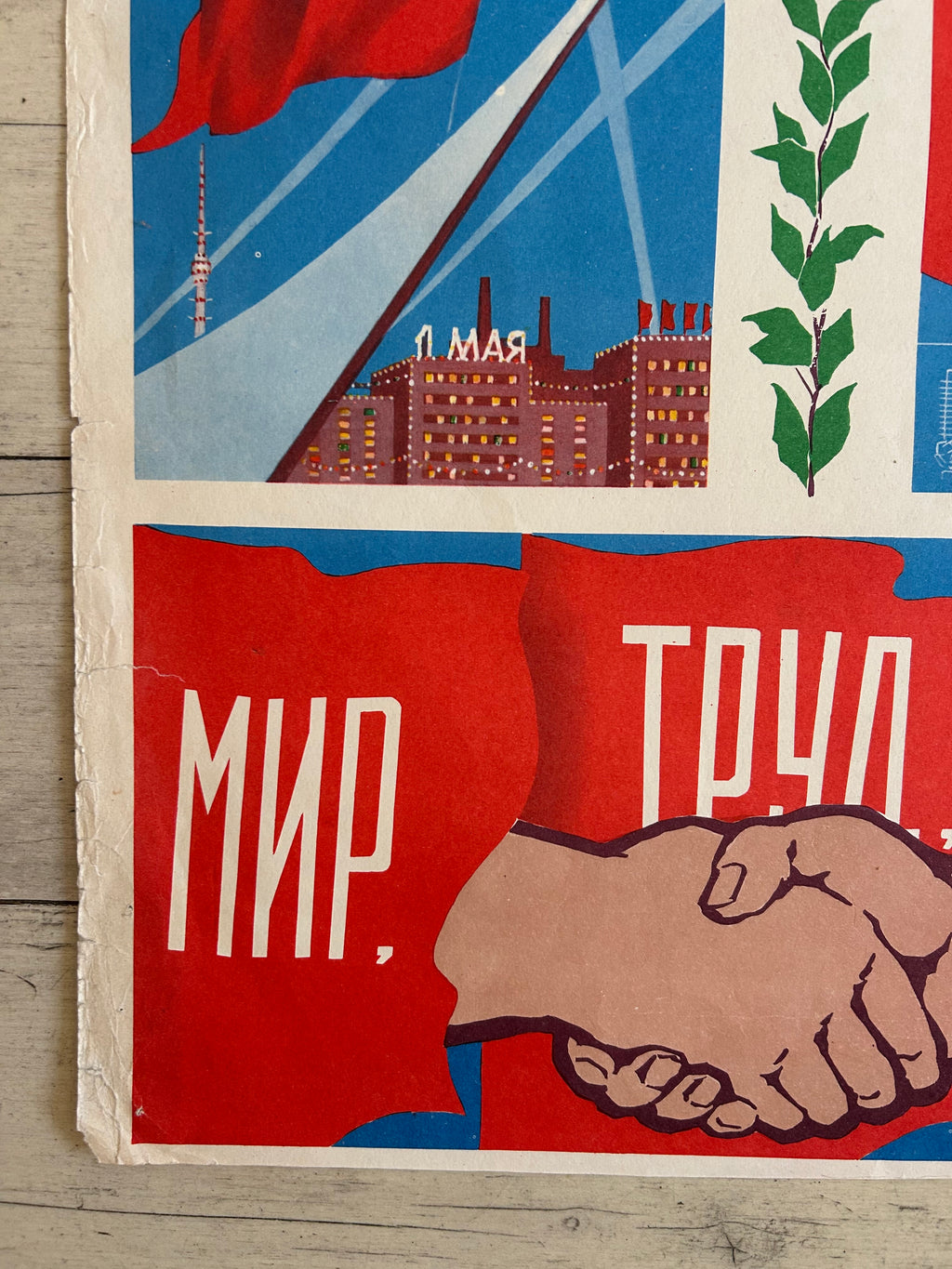 A close-up of a vintage Soviet propaganda poster featuring a handshake, a cityscape with the word 'Leningrad', a red flag, and Cyrillic text on a light wood background.
