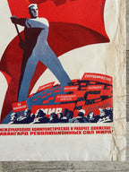 A vintage Soviet-style propaganda poster print laid flat on a weathered wood surface. The artwork features a large red flag, a blue-clad worker, and red-and-blue text blocks with Cyrillic wording.