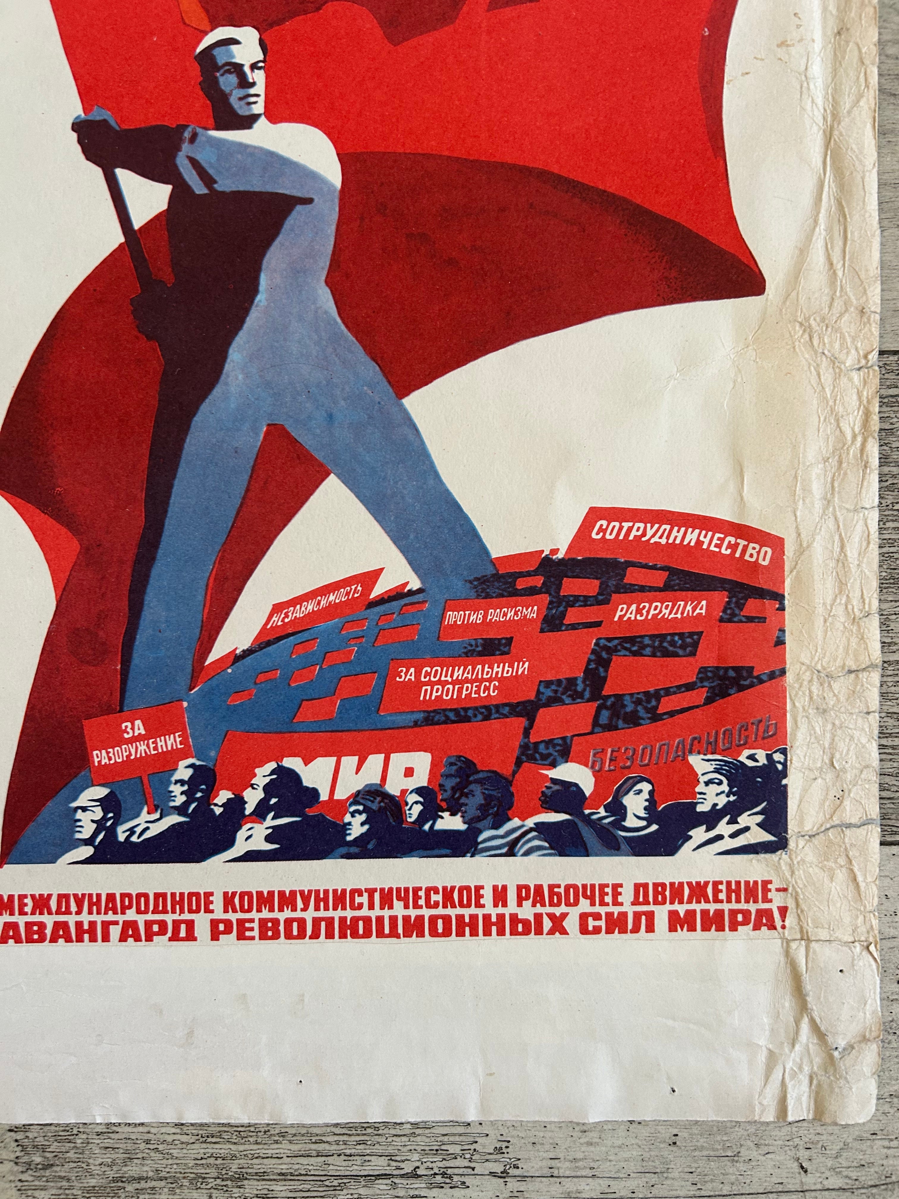 A vintage Soviet-style propaganda poster print laid flat on a weathered wood surface. The artwork features a large red flag, a blue-clad worker, and red-and-blue text blocks with Cyrillic wording.