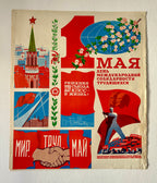 A vintage Soviet-style May Day propaganda poster with bold red, blue, and cream sections, featuring illustrations of a spire, flowers, a globe, flags, a worker, and a handshake. The Russian text reads '1 МАЯ' and other celebratory slogans.