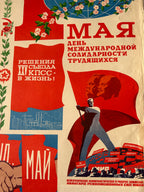 A vintage Soviet-style propaganda poster with bold red, blue, and cream sections, featuring Cyrillic text and illustrated figures with a large red hammer and sickle motif.