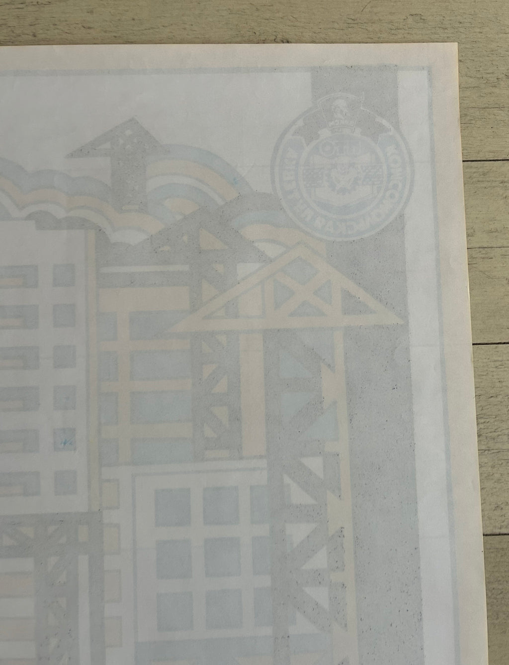 A close-up of a printed poster or art print laid on a light wood surface, showing a faint architectural illustration with a circular emblem in the upper right.