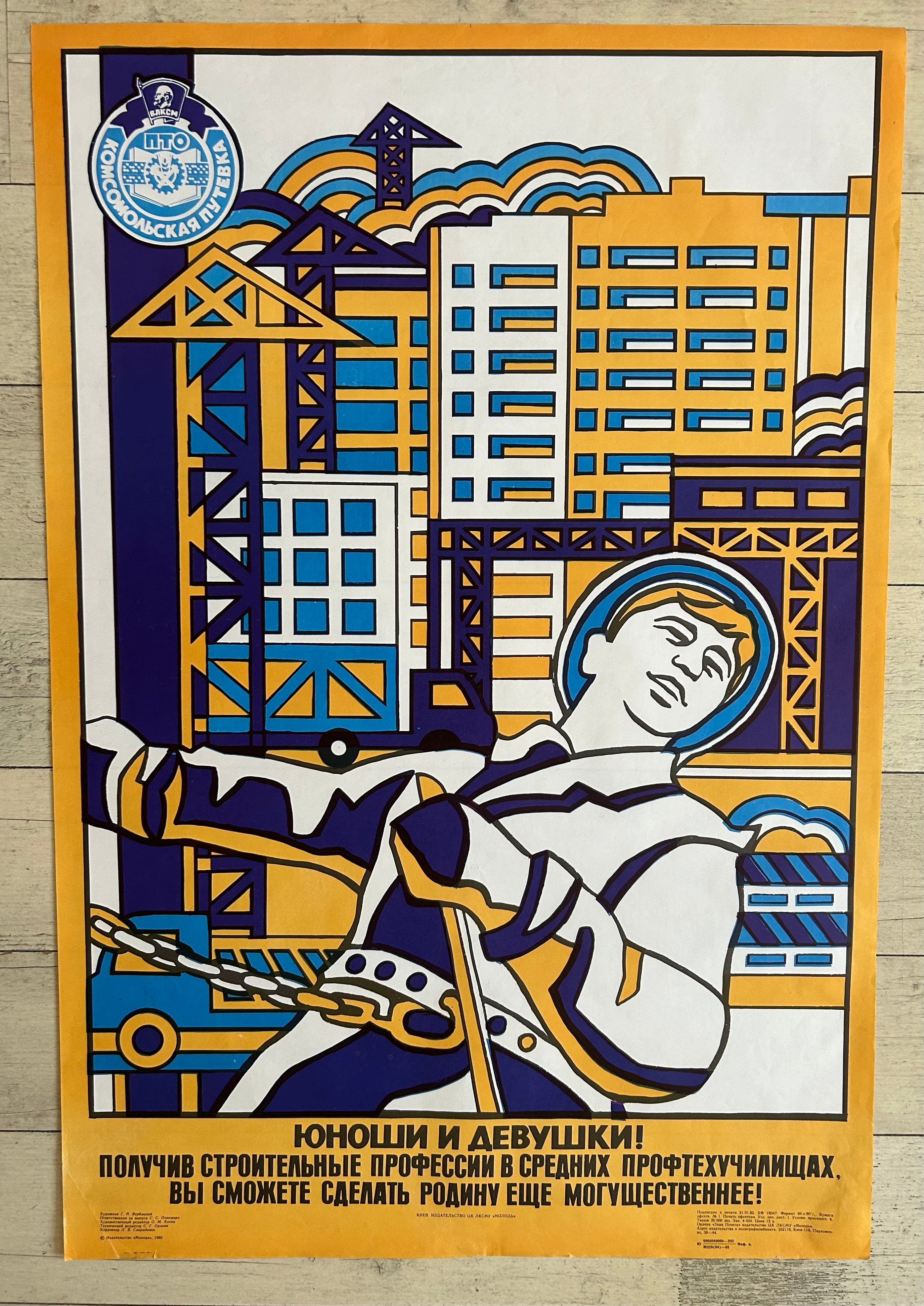 A vintage Soviet construction poster featuring a stylized worker in a white space suit pulling a chain, with cranes and buildings in blue, yellow, and white. The Russian text at the bottom reads: 'YOU HAVE RECEIVED technical training in professional construction, and you will be able to make the motherland stronger!' The poster is photographed flat against a light wood background.