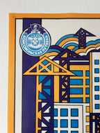 A close-up of a colorful graphic print or poster featuring stylized buildings, cranes, and a circular emblem with Cyrillic text. The artwork uses blue, yellow, purple, orange, and white in a bold geometric design, mounted against a light wall.