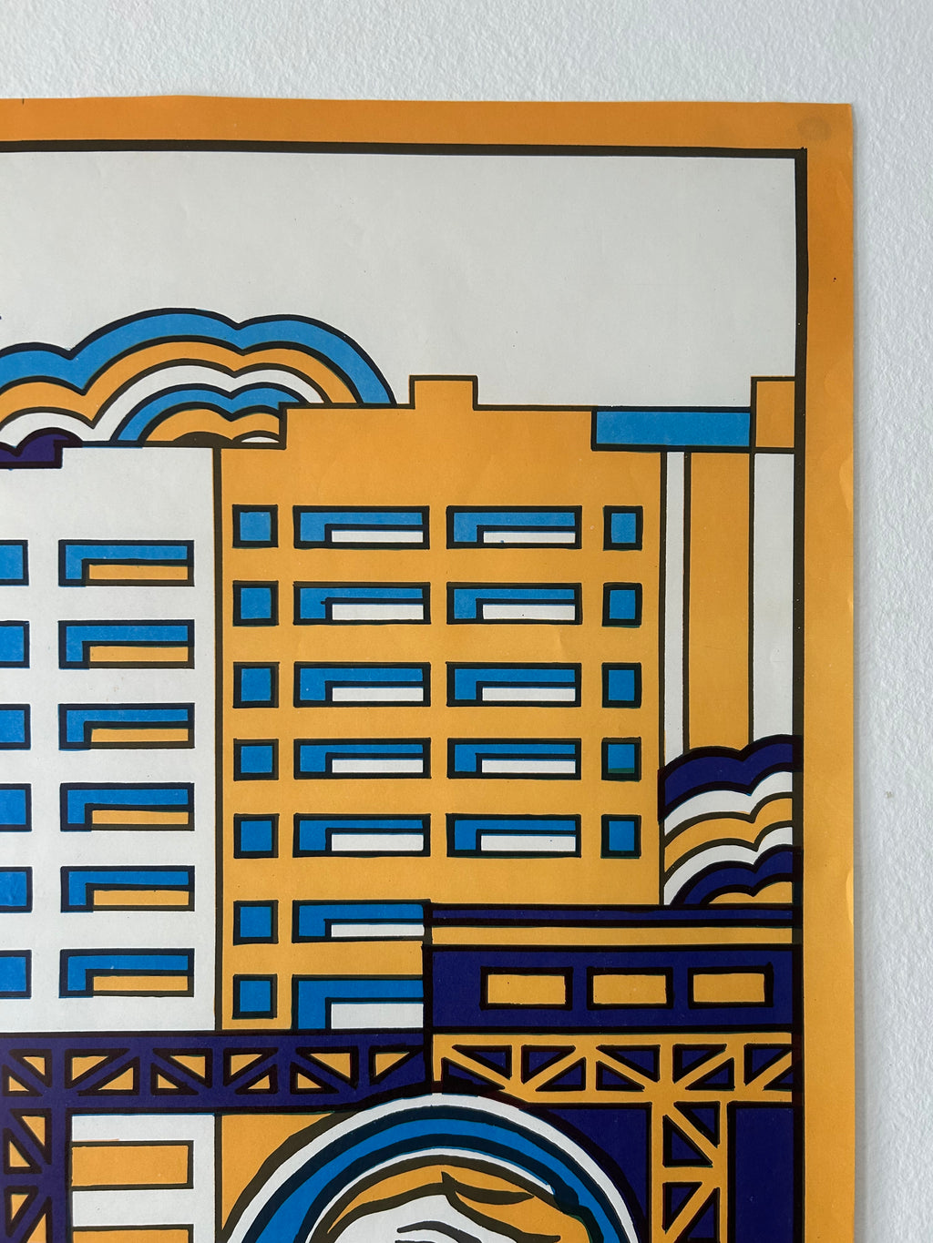 A close-up of a stylized cityscape art print featuring geometric buildings in yellow, blue, white, and dark navy, bordered by a thick orange margin, mounted on a white textured wall.