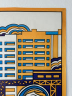 A close-up of a stylized cityscape art print featuring geometric buildings in yellow, blue, white, and dark navy, bordered by a thick orange margin, mounted on a white textured wall.