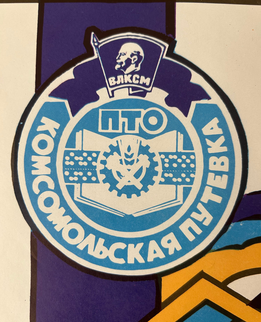 A close-up of a circular Soviet youth emblem in blue, white, and purple, featuring Cyrillic text and a central gear-and-sickle symbol.
