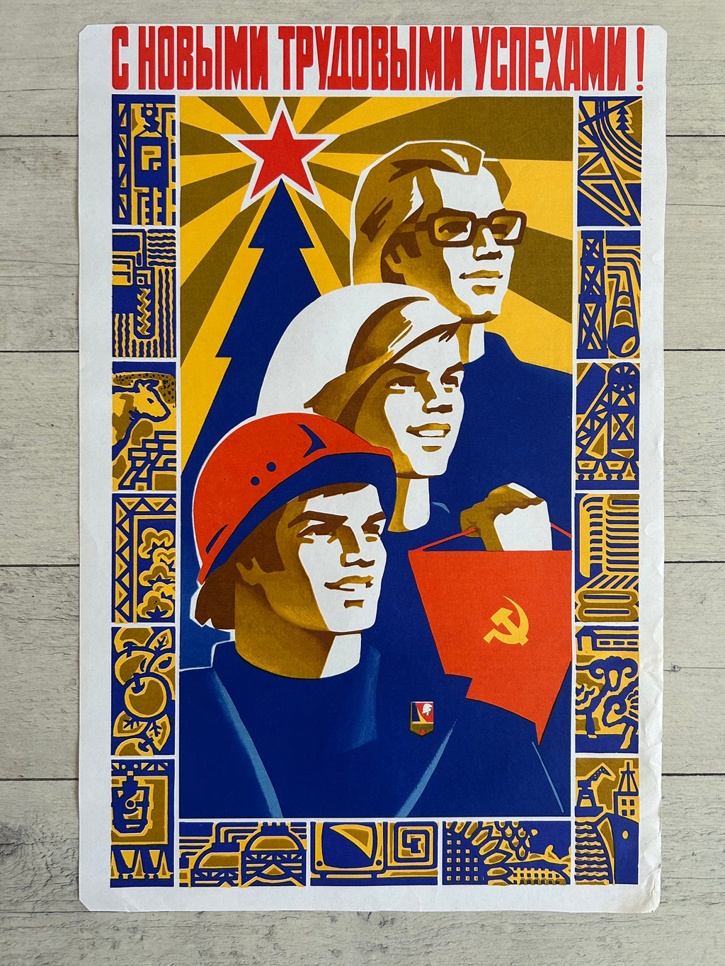 A vintage Soviet propaganda poster print featuring three workers with red stars, hats, and flags, bordered by blue and gold patterns, displayed on a light gray wood background.