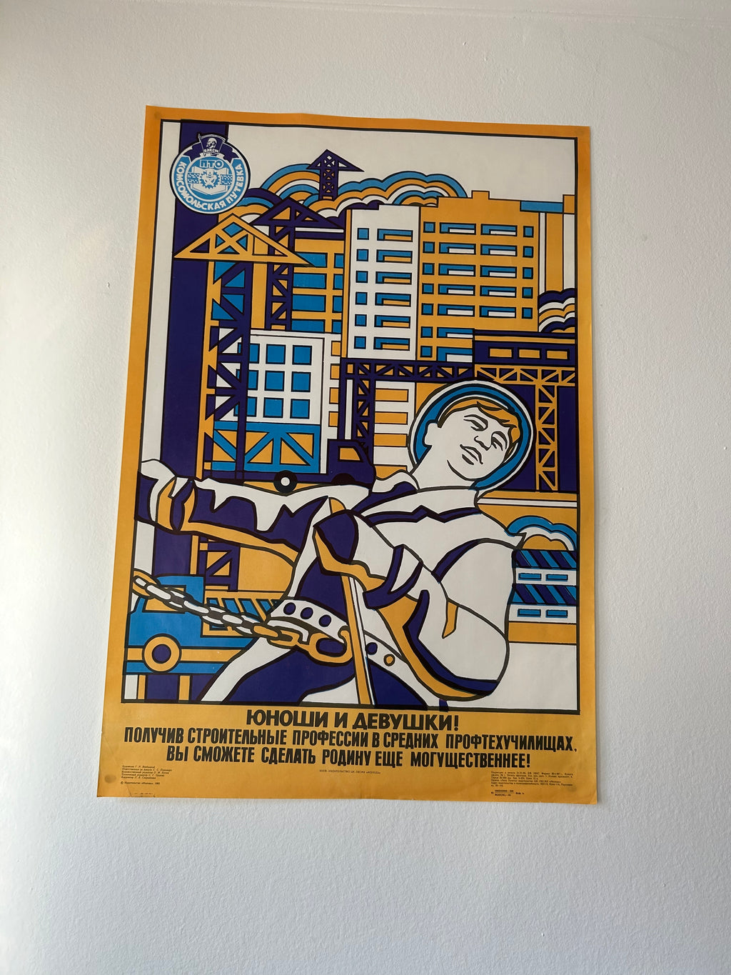 A framed Soviet-style poster mounted on a white wall, featuring a stylized construction worker in a space suit with Russian text and blue, yellow, and white graphic elements.