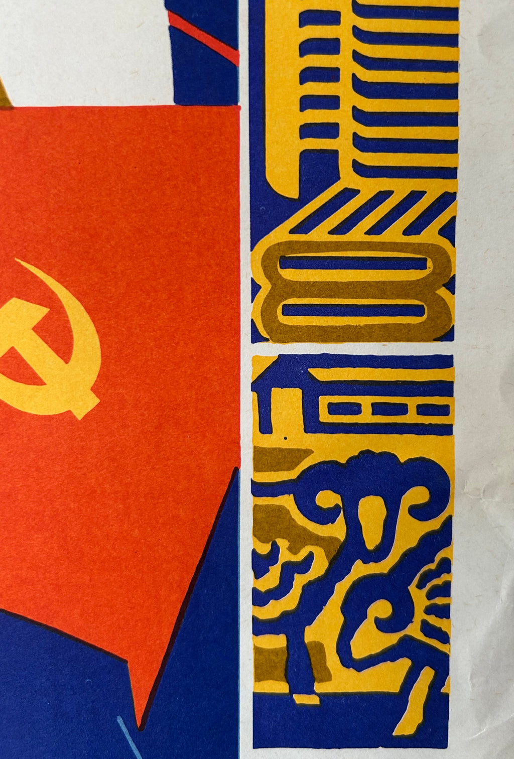 A close-up of a graphic print or poster featuring a red field with a yellow hammer and sickle symbol on the left, and a vertical column of blue and yellow geometric and decorative motifs on the right.