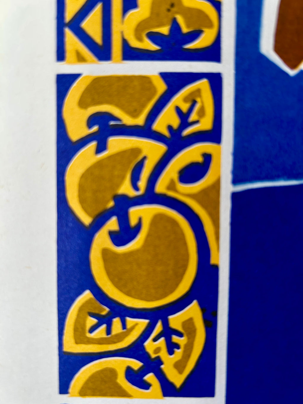 A close-up of a decorative printed pattern featuring abstract leaf or vine shapes in mustard yellow over a vivid blue background, with part of a white border and a brown shape visible at the right edge.