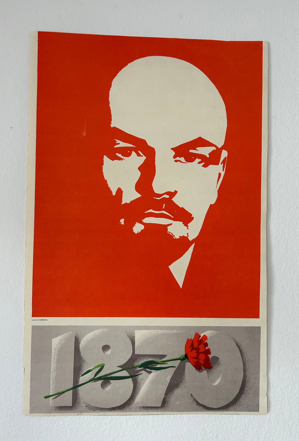 A vintage-style poster print featuring a white stencil portrait of Vladimir Lenin on a red background, with the number 1870 in gray at the bottom and a small red flower resting on the print. The poster is photographed against a white wall.