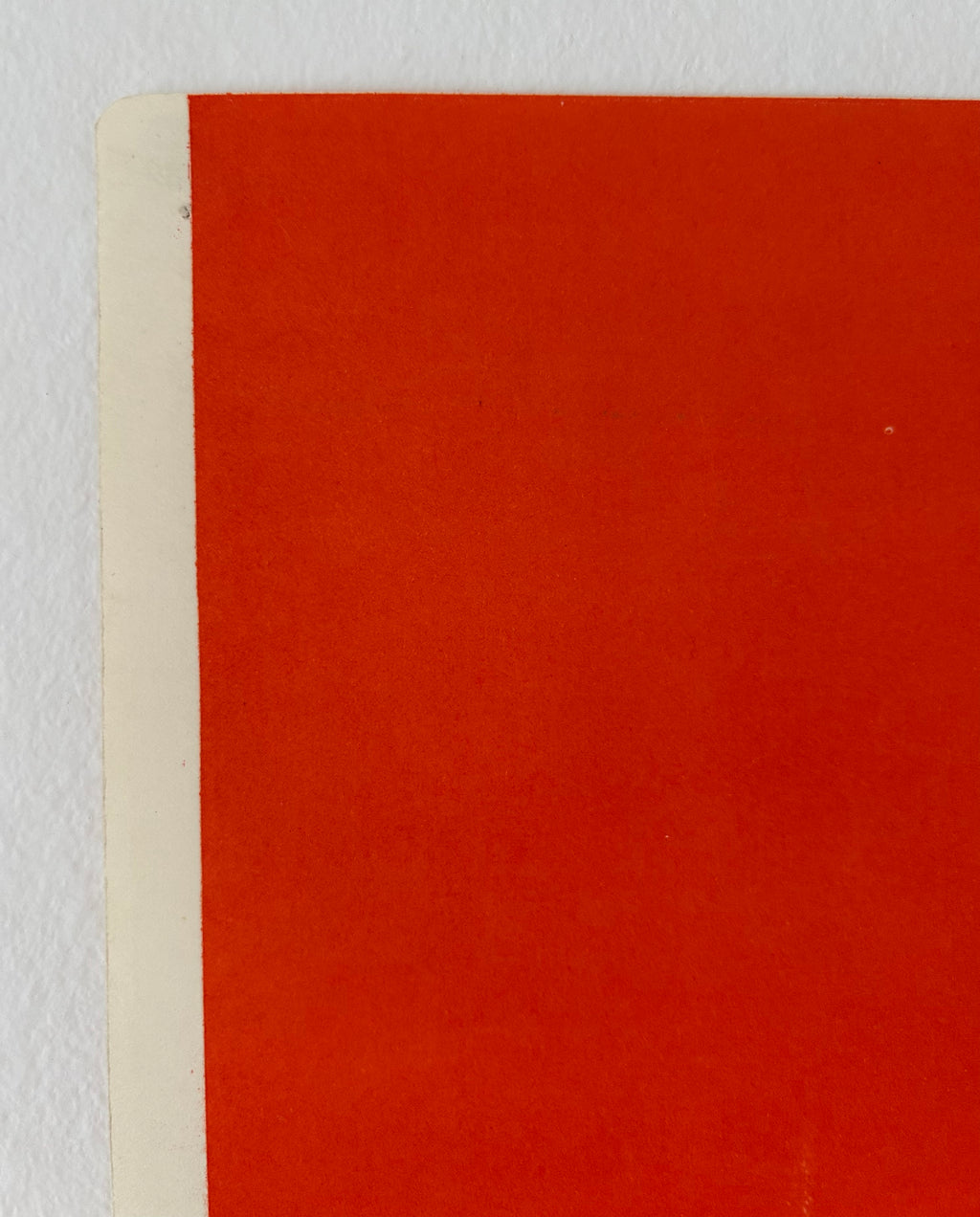 A close-up of a flat red-orange surface with a narrow off-white edge visible along the left side, photographed against a light neutral background.