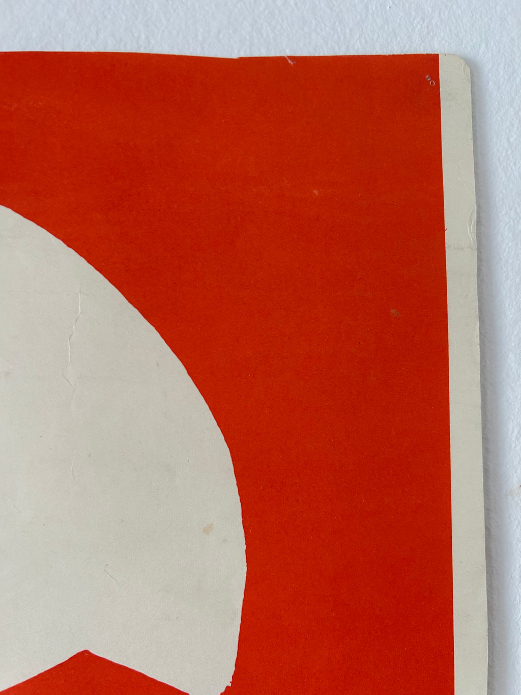 A close-up detail of a graphic art print or poster featuring a bold red background and a large off-white curved shape, with the corner of the paper visible against a light gray wall.