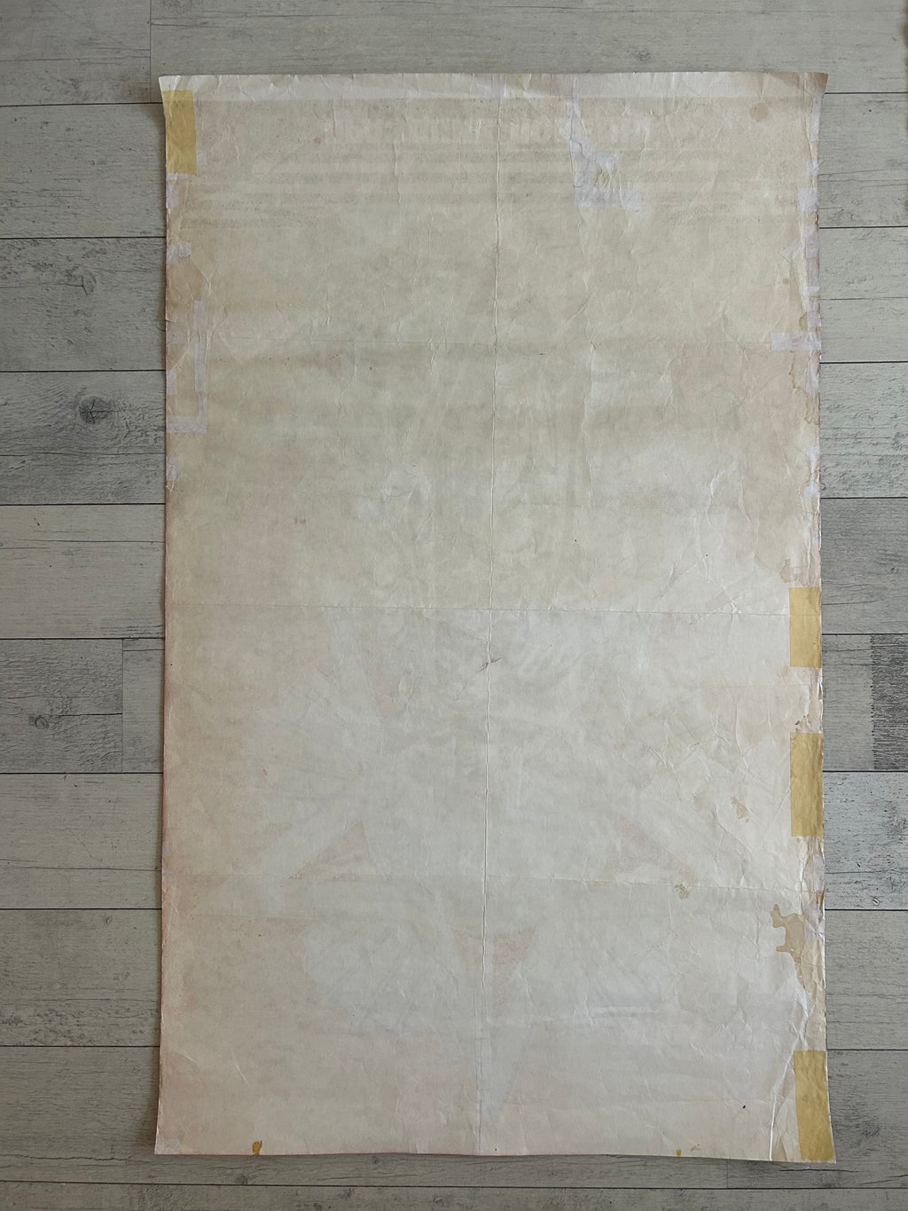 A large, flat sheet of textured beige paper or fabric with yellow tape secured along the edges, laid flat on a gray wood plank floor.