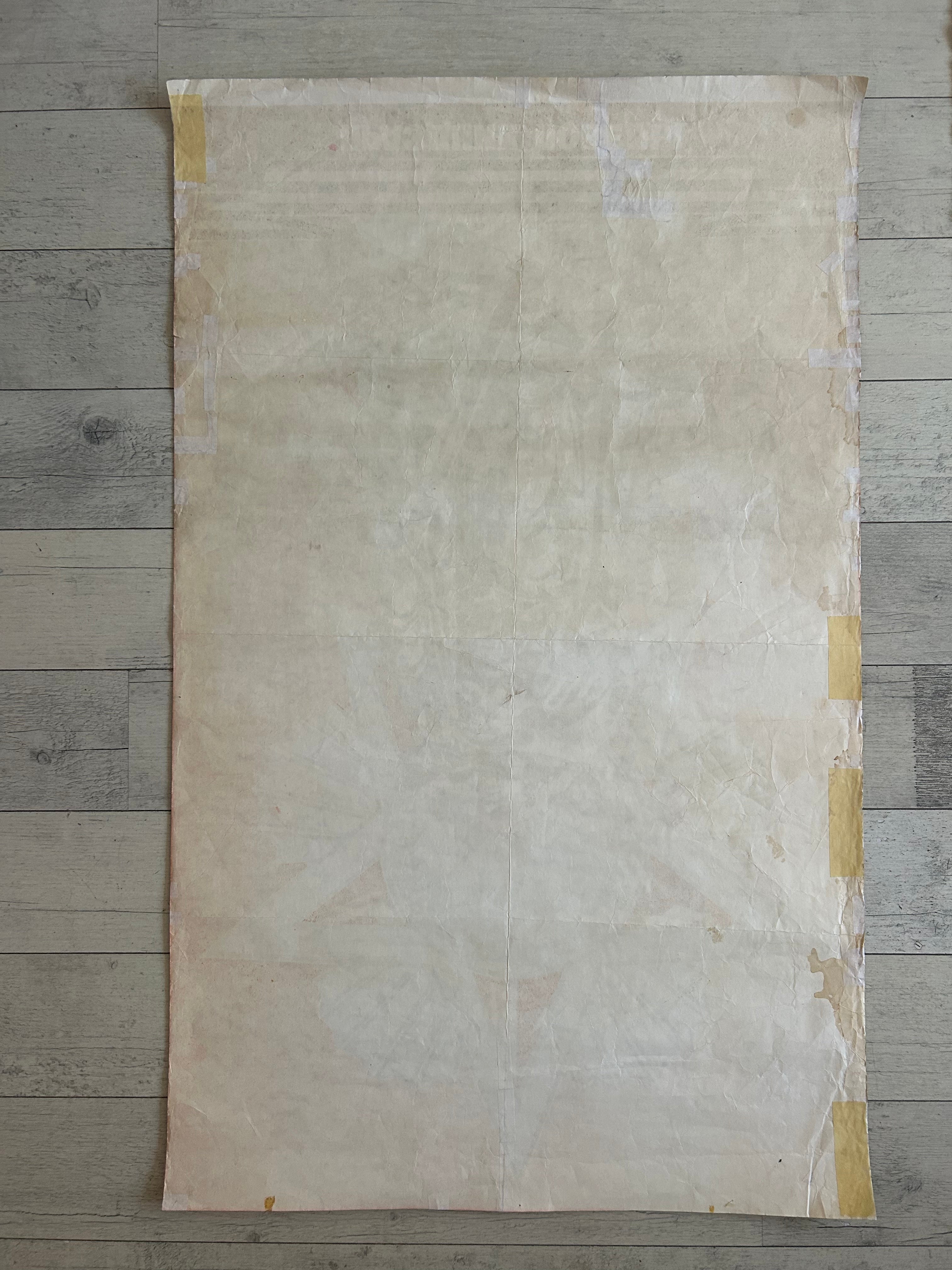 A large, flat sheet of textured beige paper or fabric with yellow tape secured along the edges, laid flat on a gray wood plank floor.
